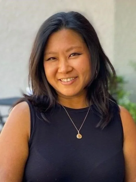 A woman with shoulder-length black hair, smiling, wearing a sleeveless black top and a gold necklace with a round pendant, standing outdoors.