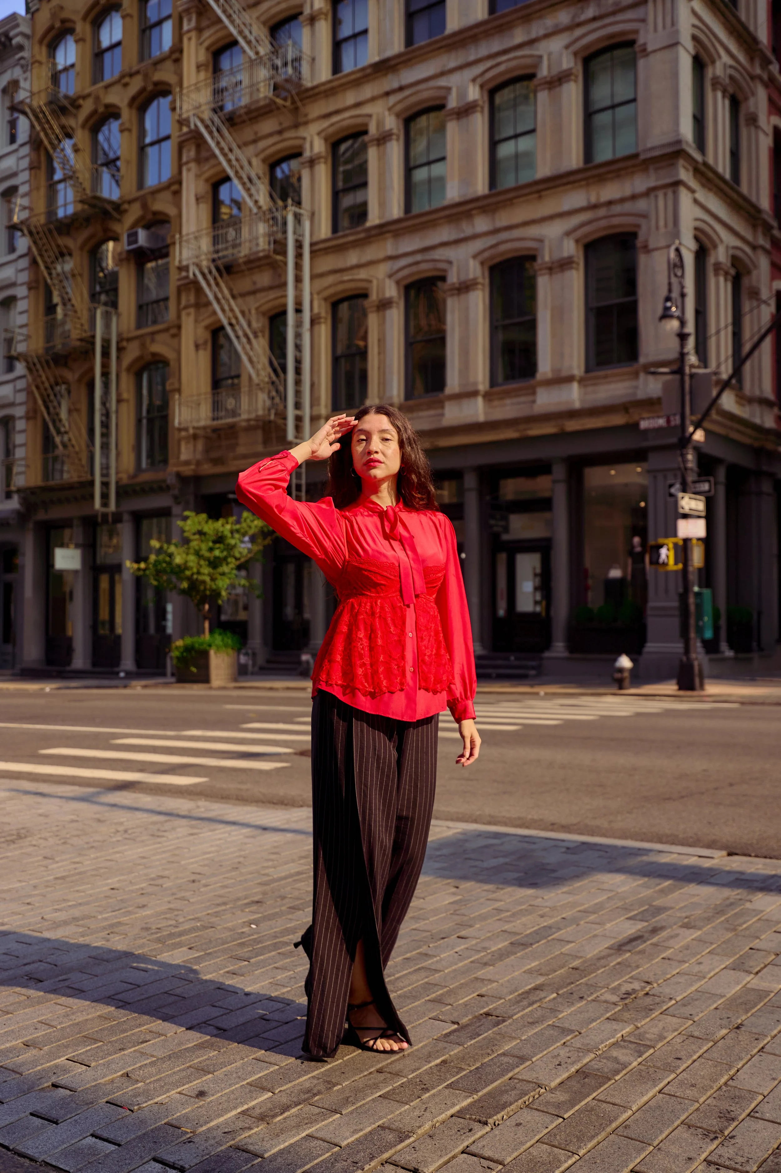 A woman with dark curly hair standing on a city street at sunset, wearing a red blouse with lace details and black pinstripe pants.