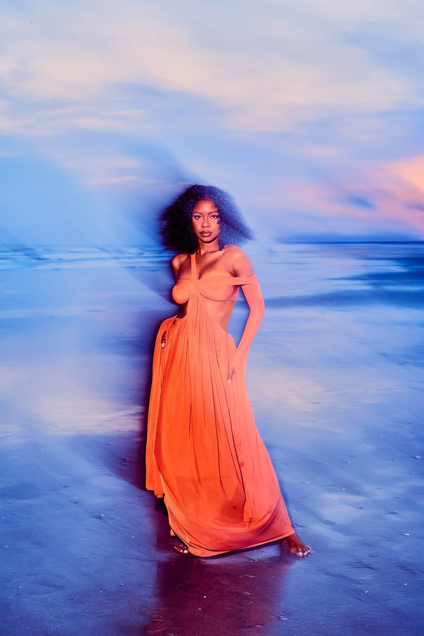 A woman with curly hair in an orange dress standing on a beach at sunset, with waves in the background.