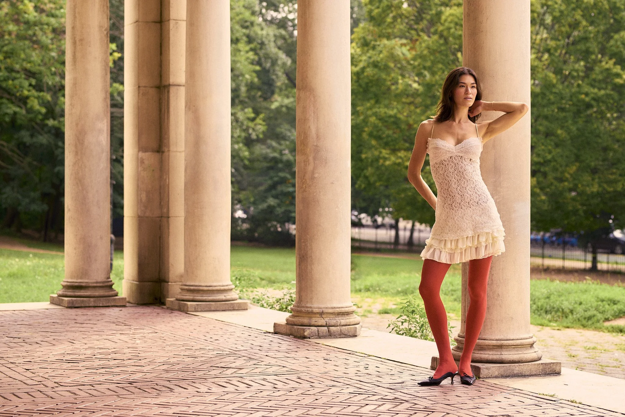 A woman in a lace dress, red tights, and high heels standing between beige stone columns in a park setting with green trees in the background.