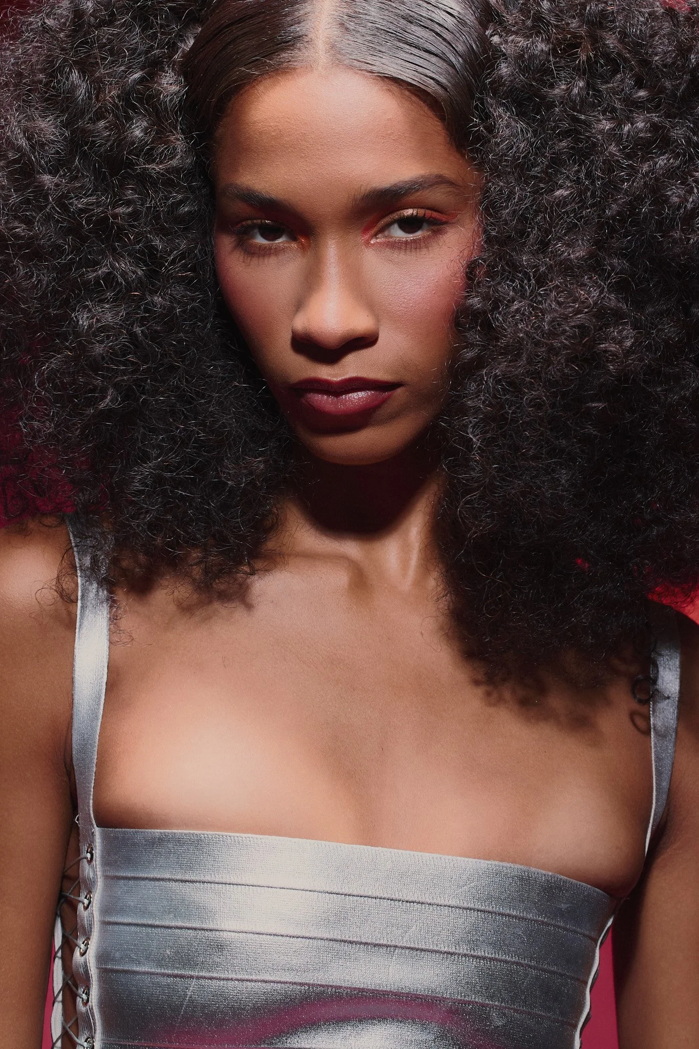 Close-up of a woman with voluminous curly hair, wearing a silver satin top with lace-up sides, looking directly at the camera with a confident expression.