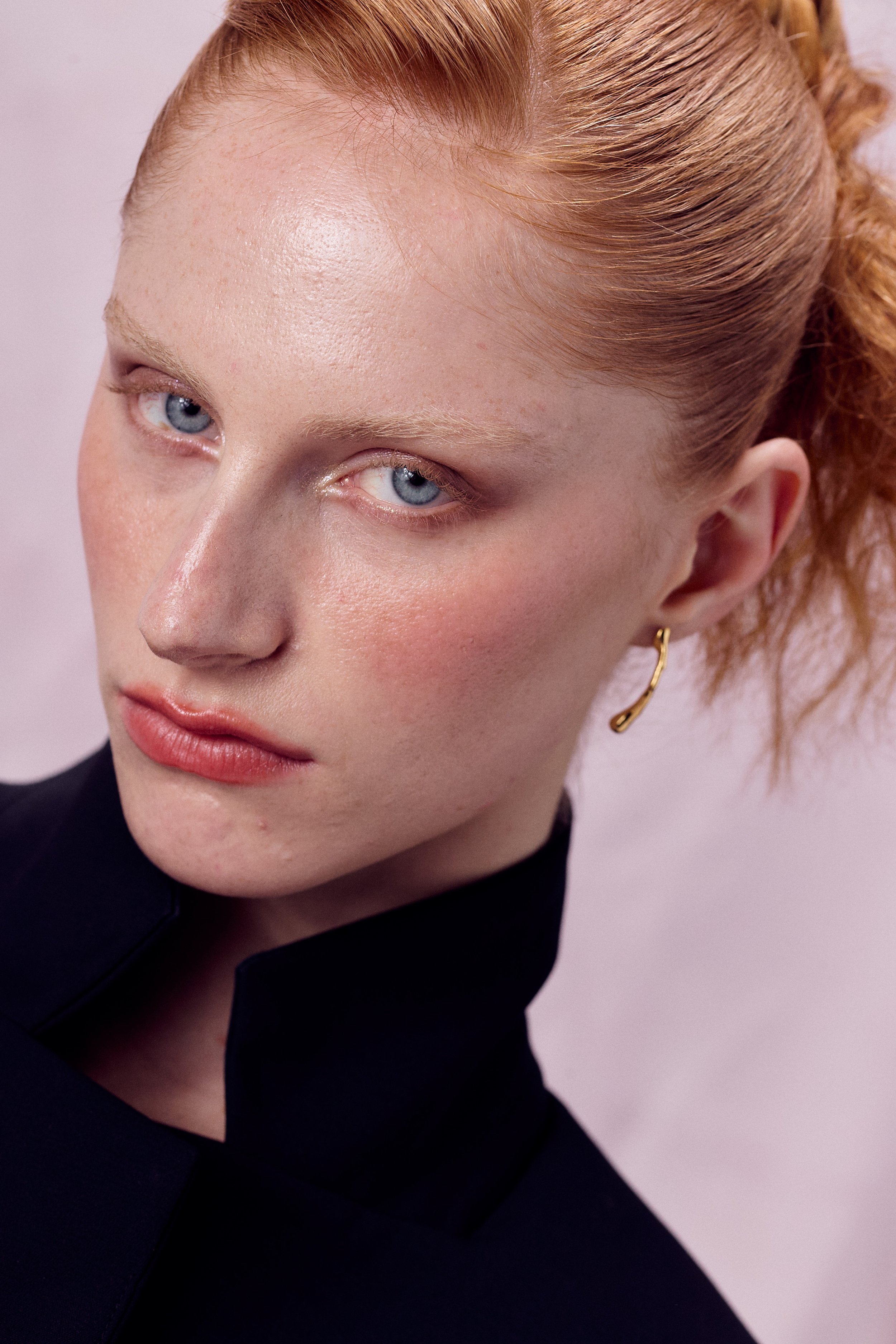 Close-up of a woman's face with red hair, blue eyes, light makeup, and a gold hoop earring, wearing a dark top, against a light pink background.