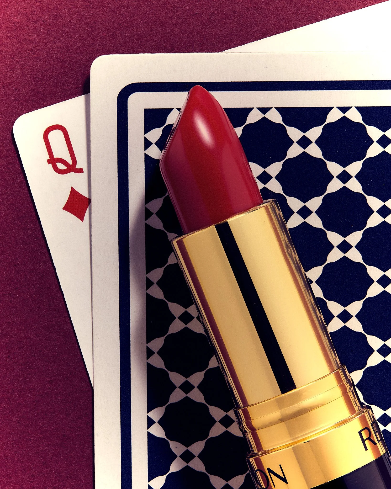 Close-up of a red lipstick tube with a gold band, resting on two playing cards with a patterned background and maroon fabric.