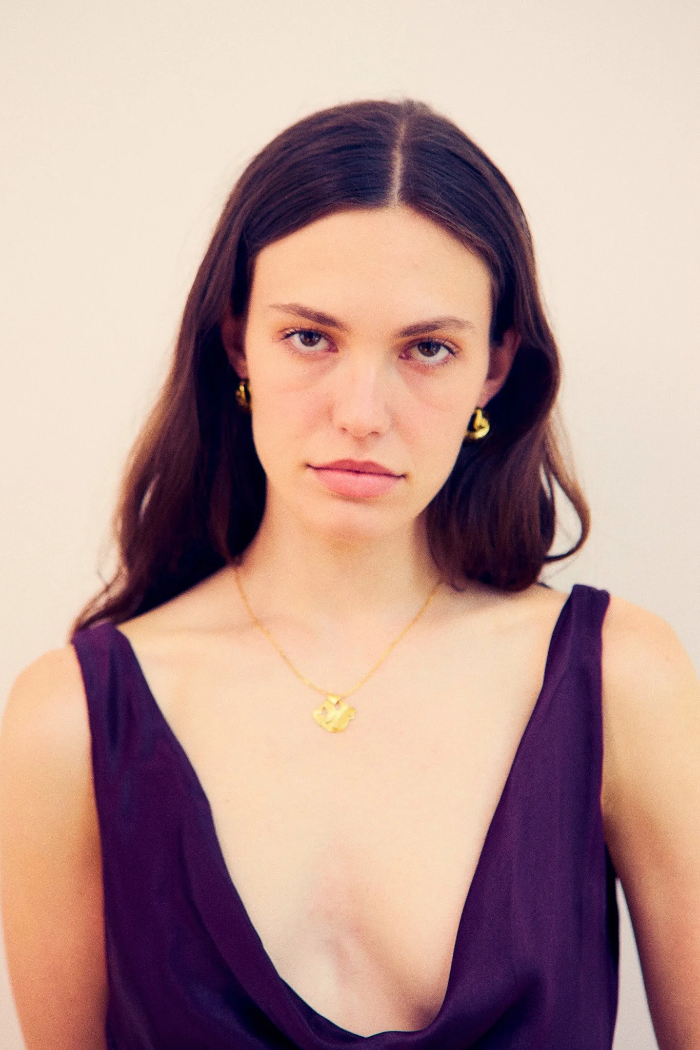 A young woman with long dark brown hair styled in loose waves, wearing a purple sleeveless dress with a deep neckline, gold hoop earrings, and a gold necklace with a pendant featuring a floral design, against a plain white background.