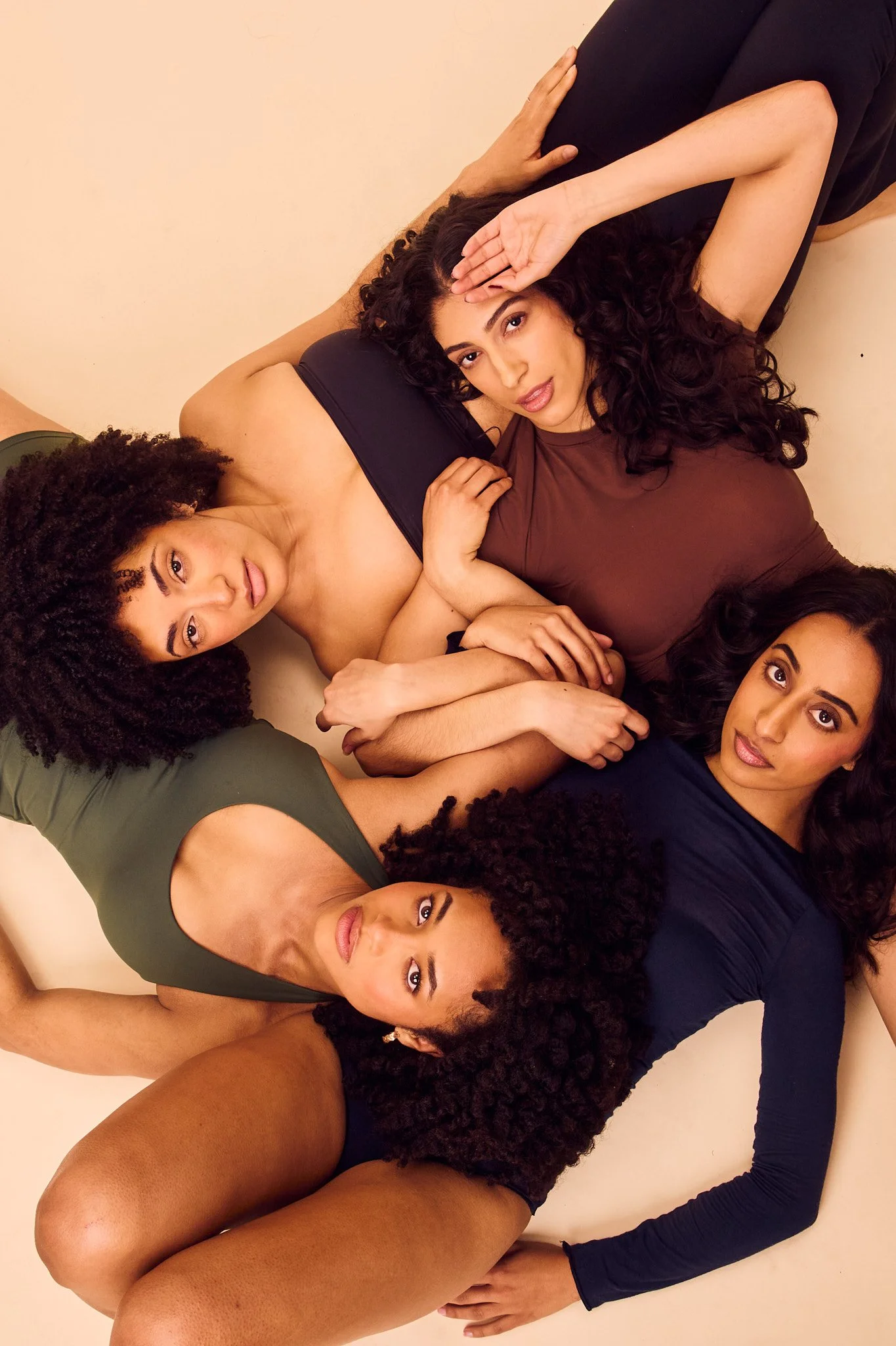 Six women with various skin tones and curly hair lying on the floor in a circle, facing upward, with their heads close together and arms around each other.