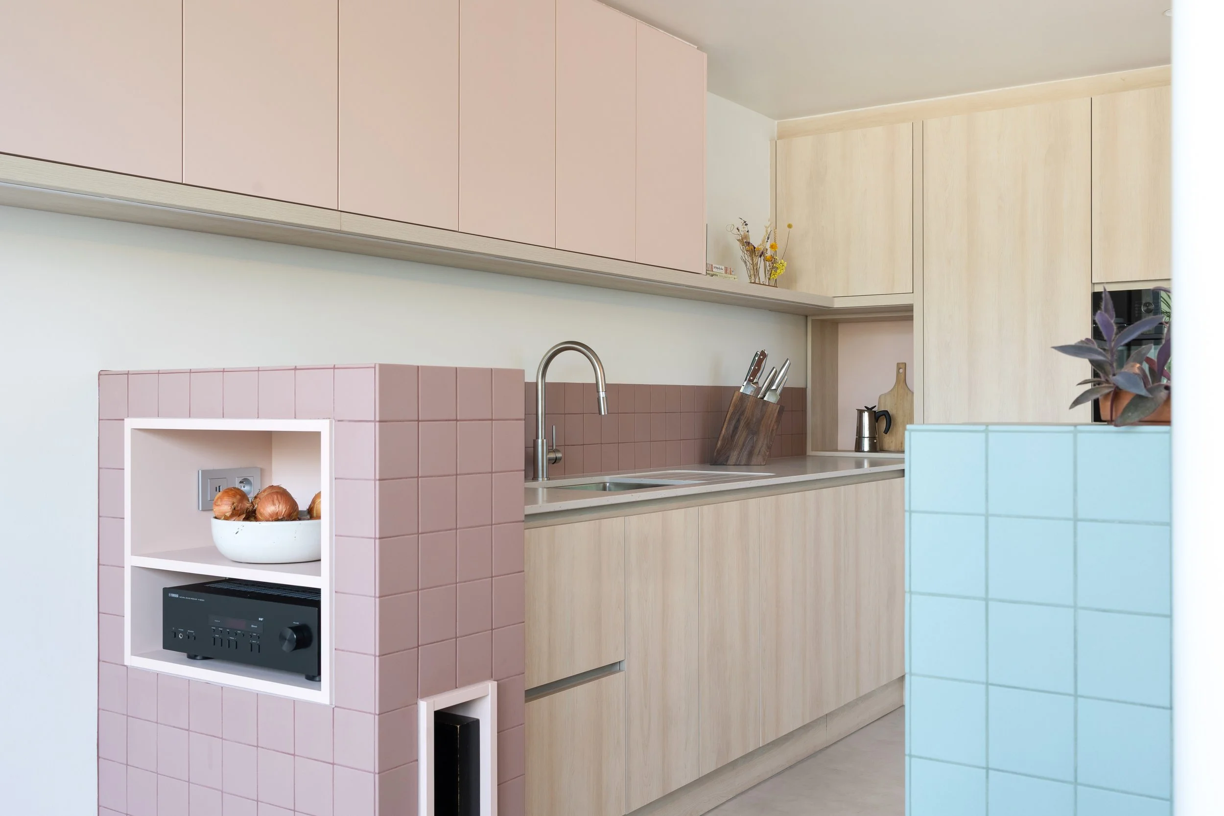 Keuken met roze tegels achter het aanrecht, lichte houten kasten en een roze betegelde kast met open nis