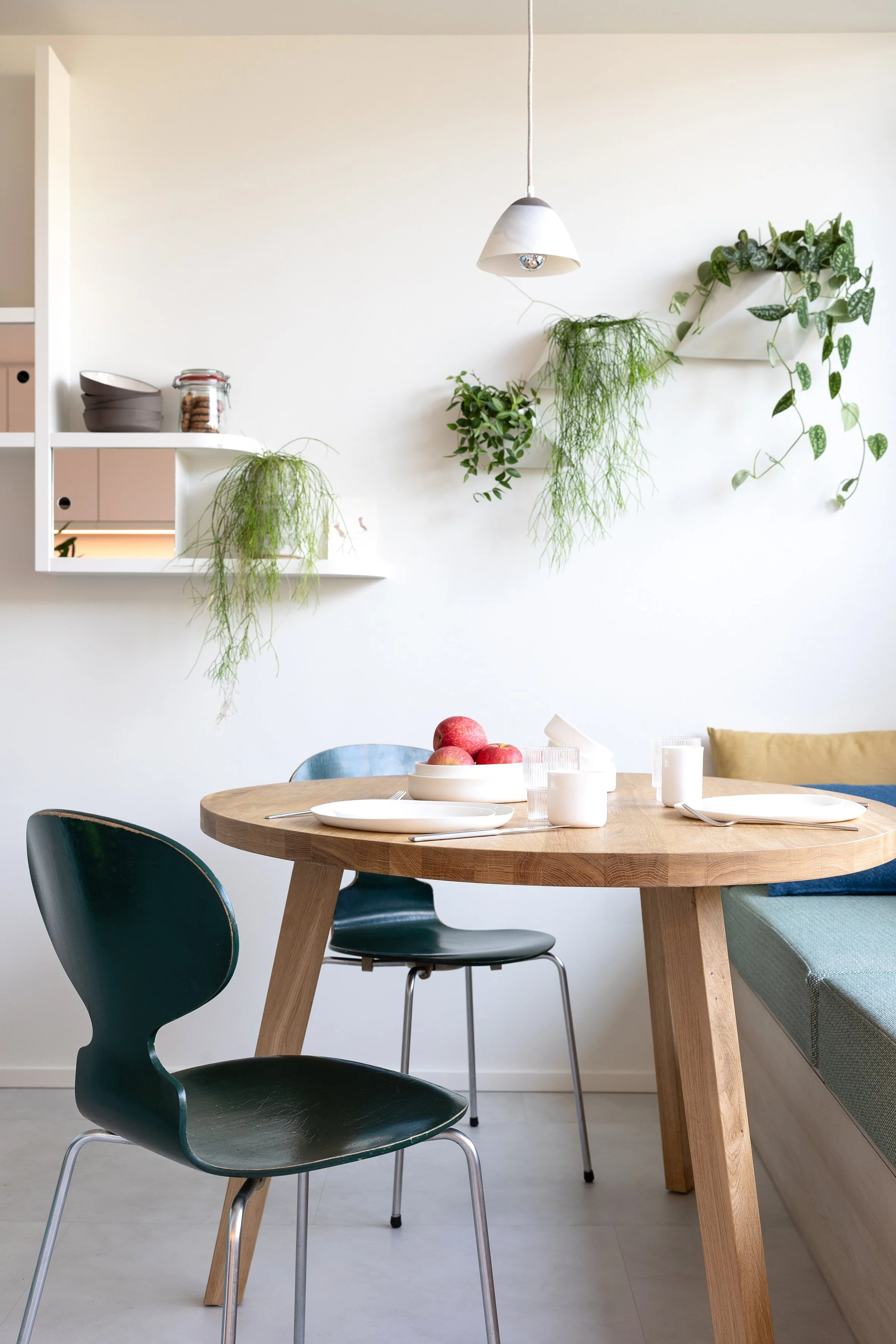 Eethoek met gerecupereerde eiken tafel, donkergroene stoelen en hangplanten aan een witte wand