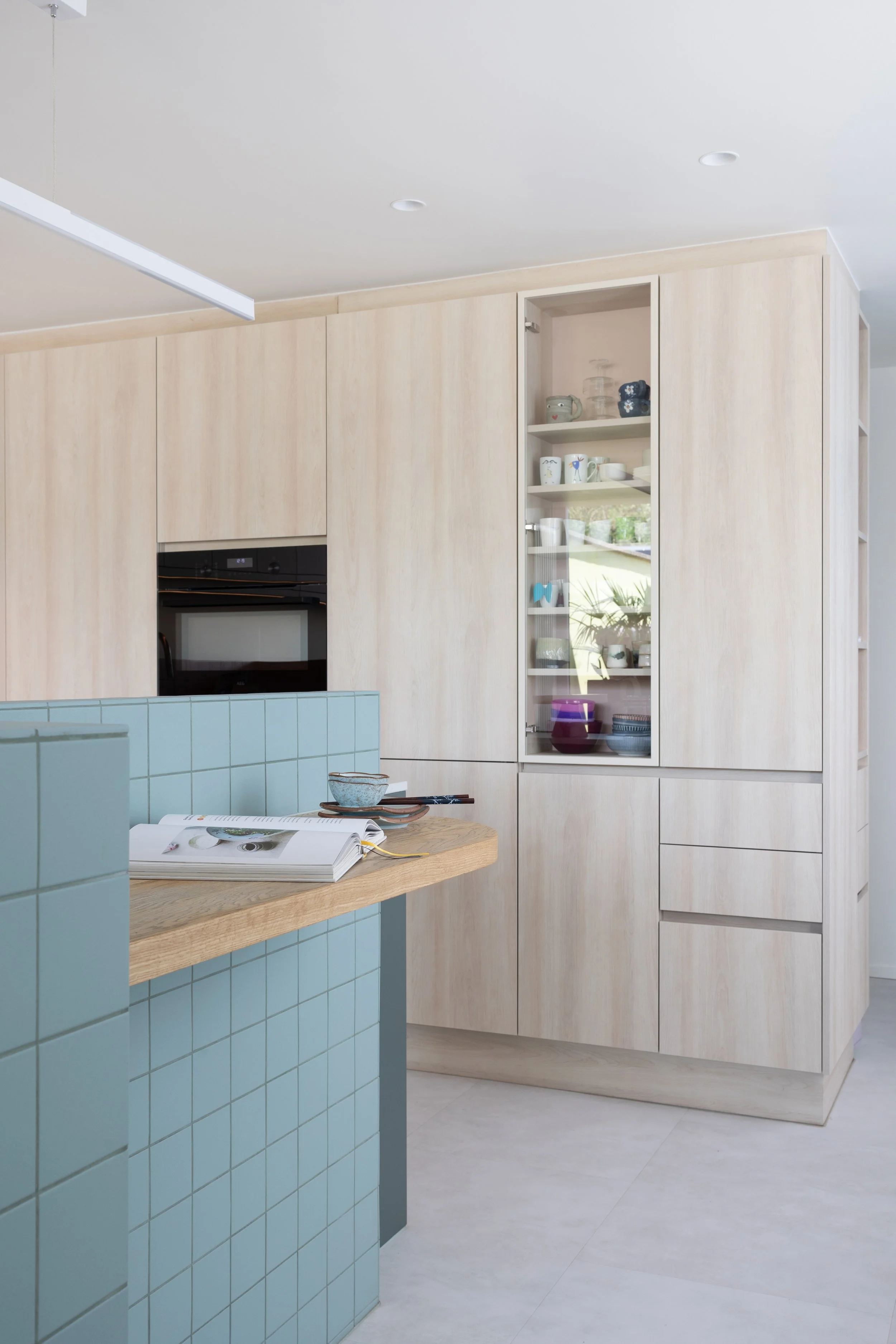 Kastenwand in lichte houtstructuur met oven en glazen vitrinekast, met het betegelde eiland en werktablet op de voorgrond