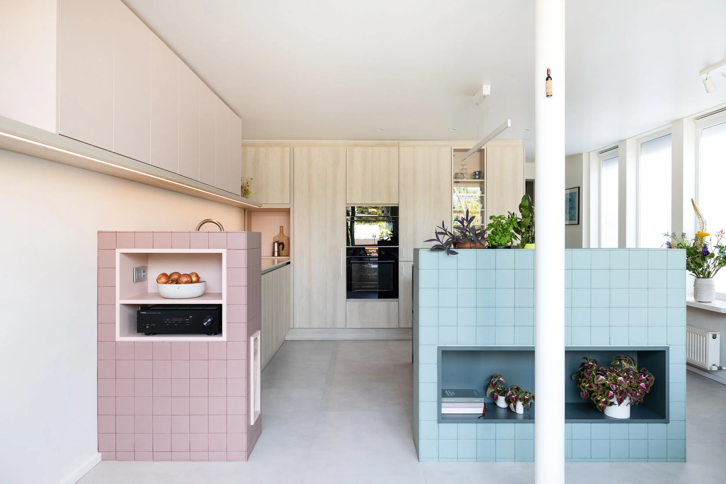 Open keuken met een roze betegelde kast met open nis en een saliegroen betegeld keukeneiland met plantennis