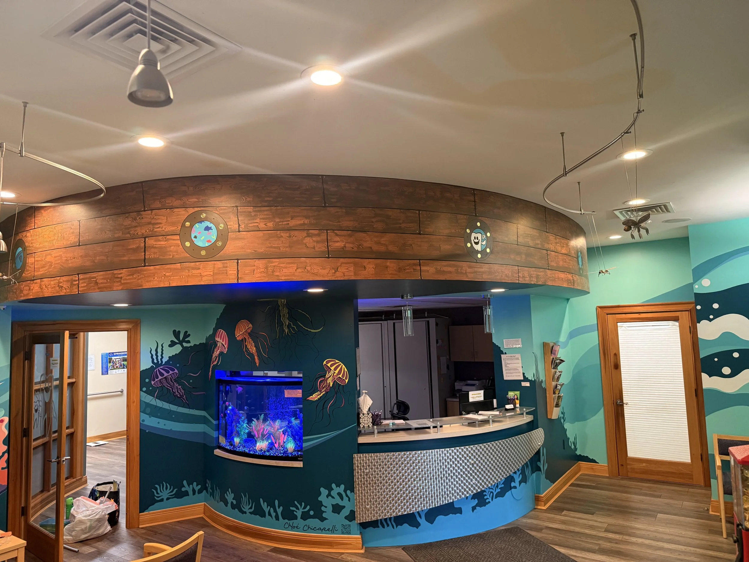 Interior of an aquarium or marine-themed room with a reception desk, colorful jellyfish wall art, an aquarium tank, and a turquoise underwater mural.