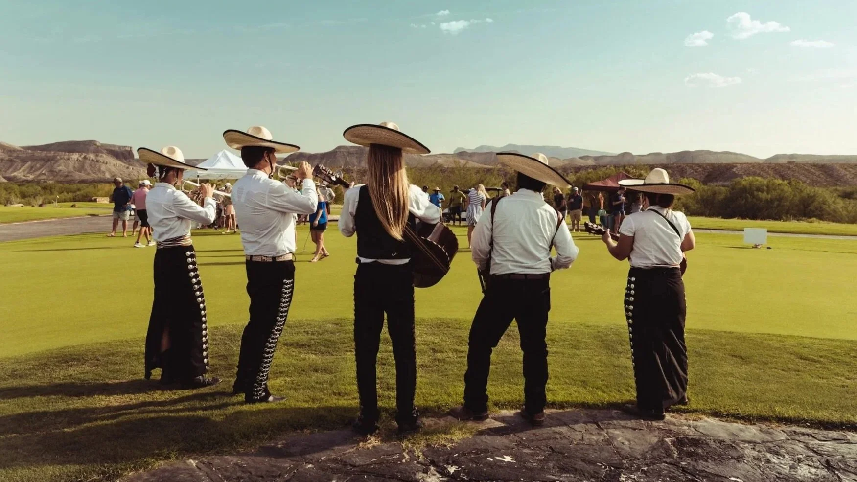 Gruppo di musicisti vestiti con abiti tradizionali messicani, alcuni che suonano strumenti come chitarre e flauti, mentre altri ascoltano in un campo da golf con paesaggi montuosi sullo sfondo.