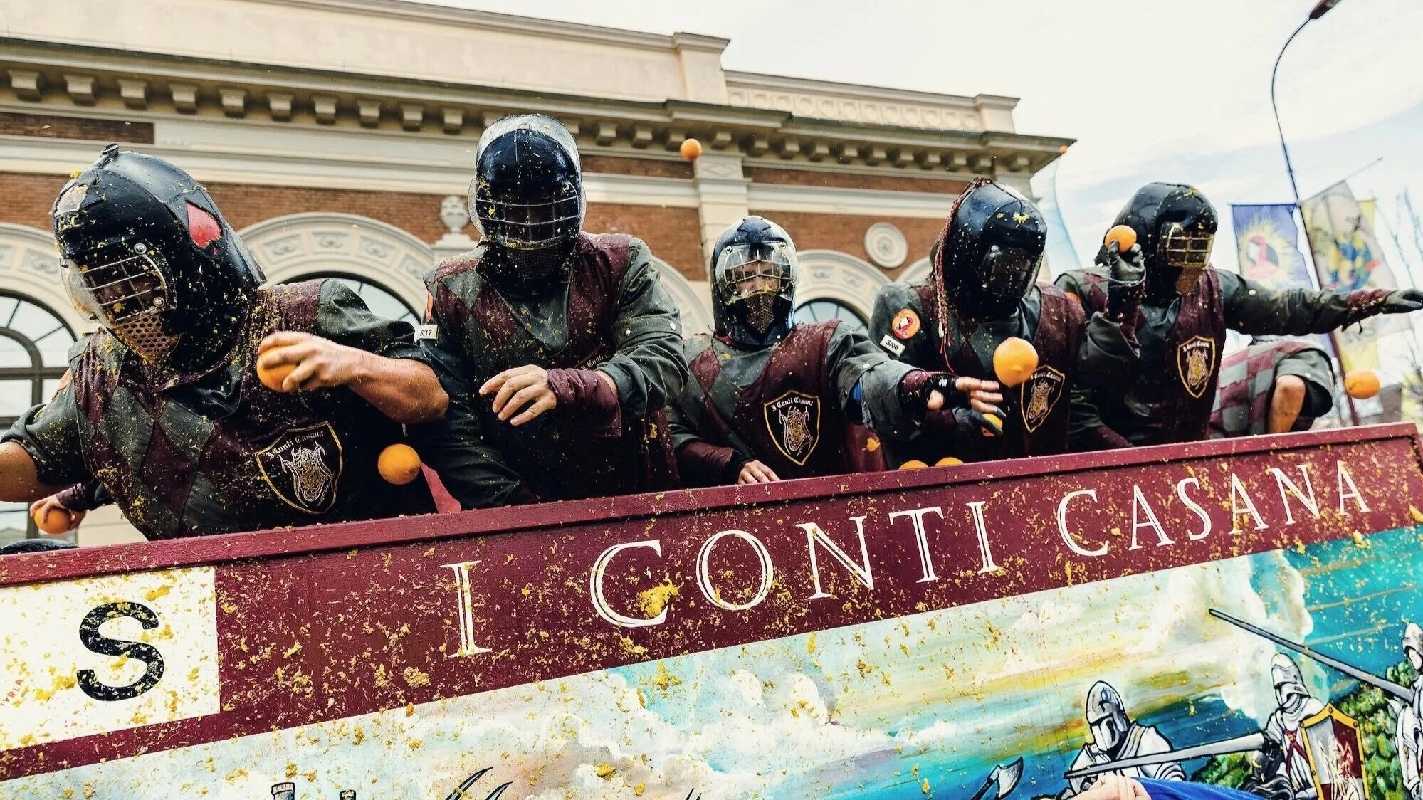 Gruppo di persone in maschera su una barca decorata durante una festa tradizionale, con un edificio storico sullo sfondo. Sono tutti vestiti con costumi scuri e caschi con visiere.