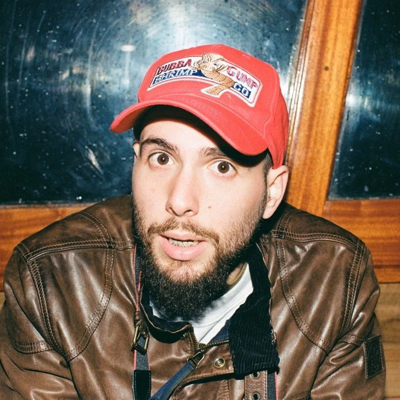 Uomo con cappello rosso e giacca di pelle marrone, con sfondo di legno e vetro