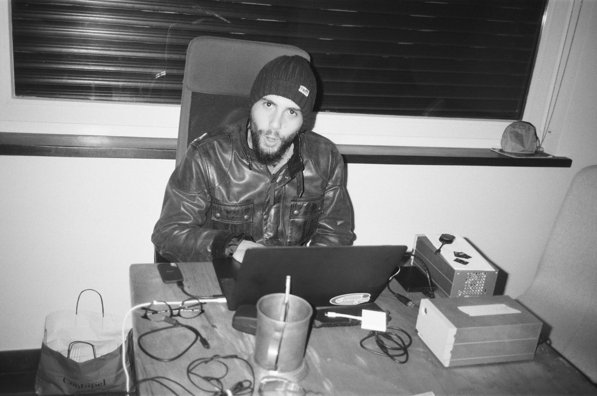 Uomo con cappello di lana e giacca in pelle seduto davanti a un computer portatile in un ufficio, con vari oggetti e cavi sul tavolo.