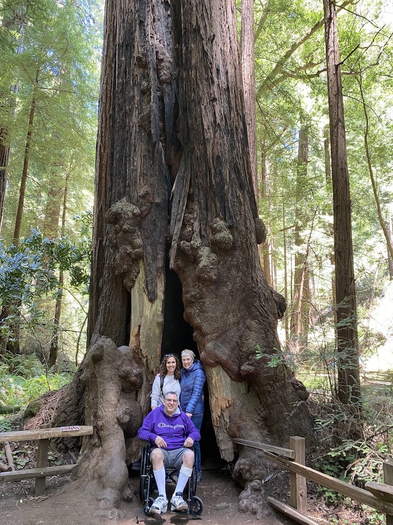 A mother, daughter and son in a wheelchair in front of a large redwood tree at Muir Woods