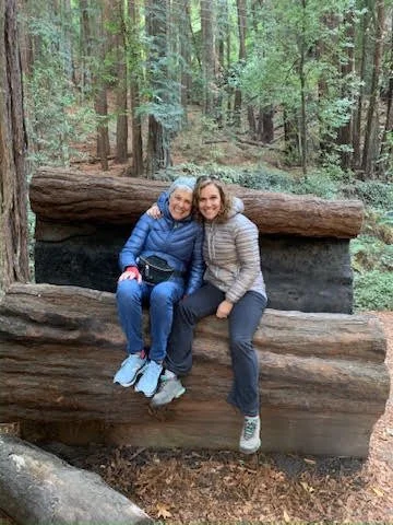 A mother and daughter sit holding each other on a bench made out of a redwood tree in a redwood forest