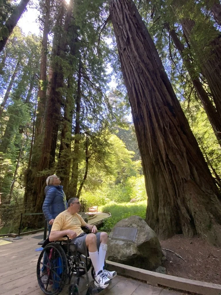 an older woman and man in a wheelchair are on a wooden walkway among the redwoods at Muir Woods