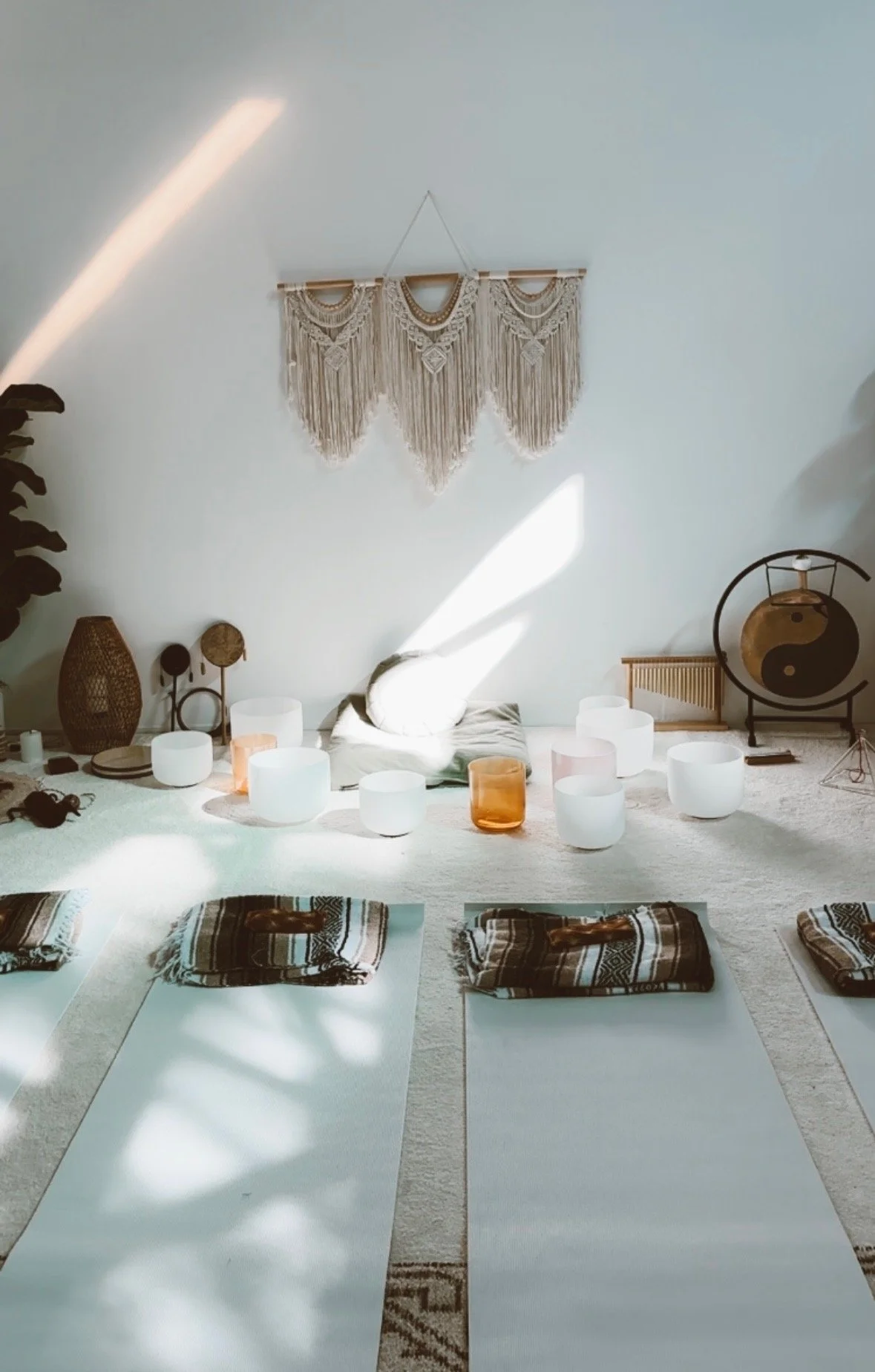 A yoga or meditation space with mats and blankets on a light-colored rug, decorated with candles, lamps, and spiritual decor like a yin-yang drum and woven wall hanging, in a room with sunlight streaming through.