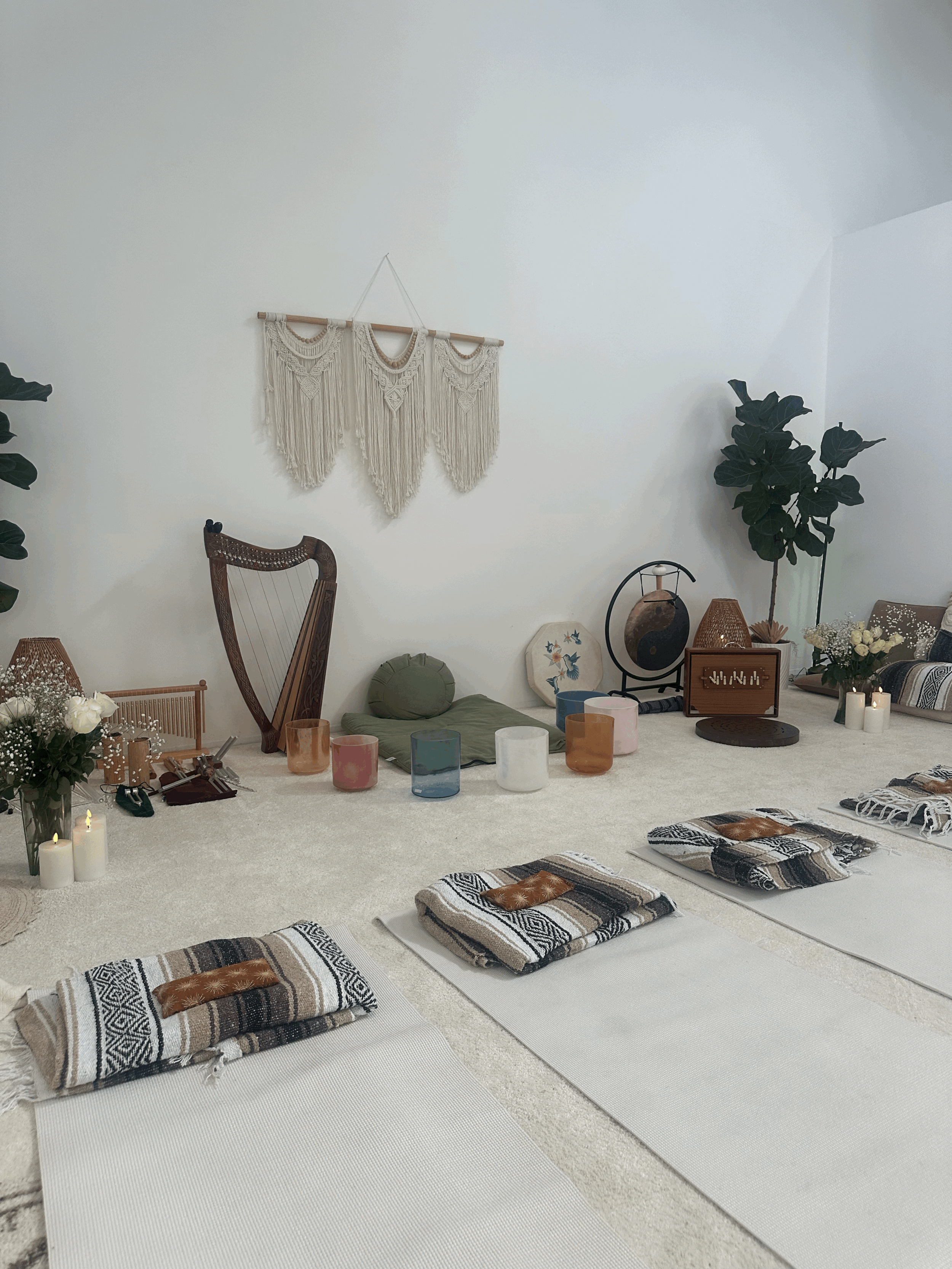 A cozy meditation or yoga space with mats and pillows on a white carpet, surrounded by candles, plants, and decorative items like a harp, vases, and artwork on the wall.