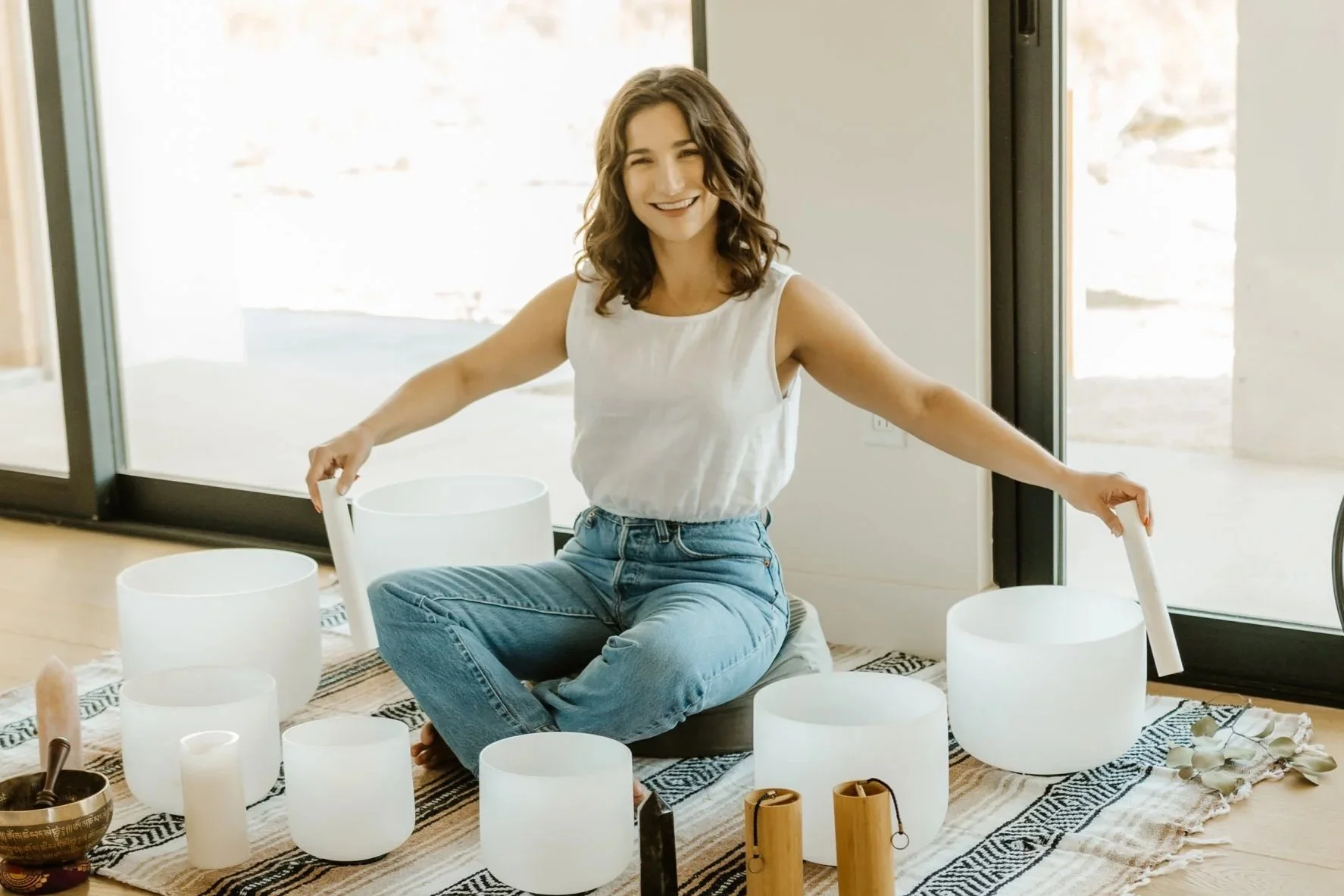 A woman sitting on a cushion on a rug inside, surrounded by quartz singing bowls and other instruments, smiling with arms extended, near a large window.
