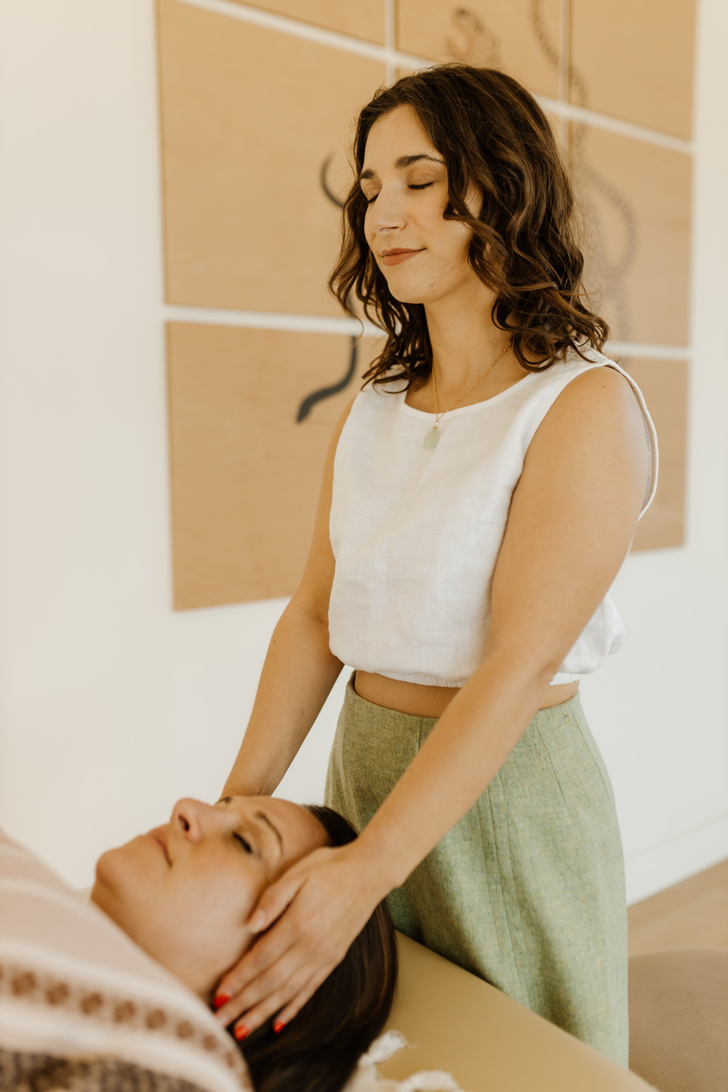 A woman with curly brown hair gently holds a woman's head as she receives a massage or scalp treatment in a wellness or spa setting.