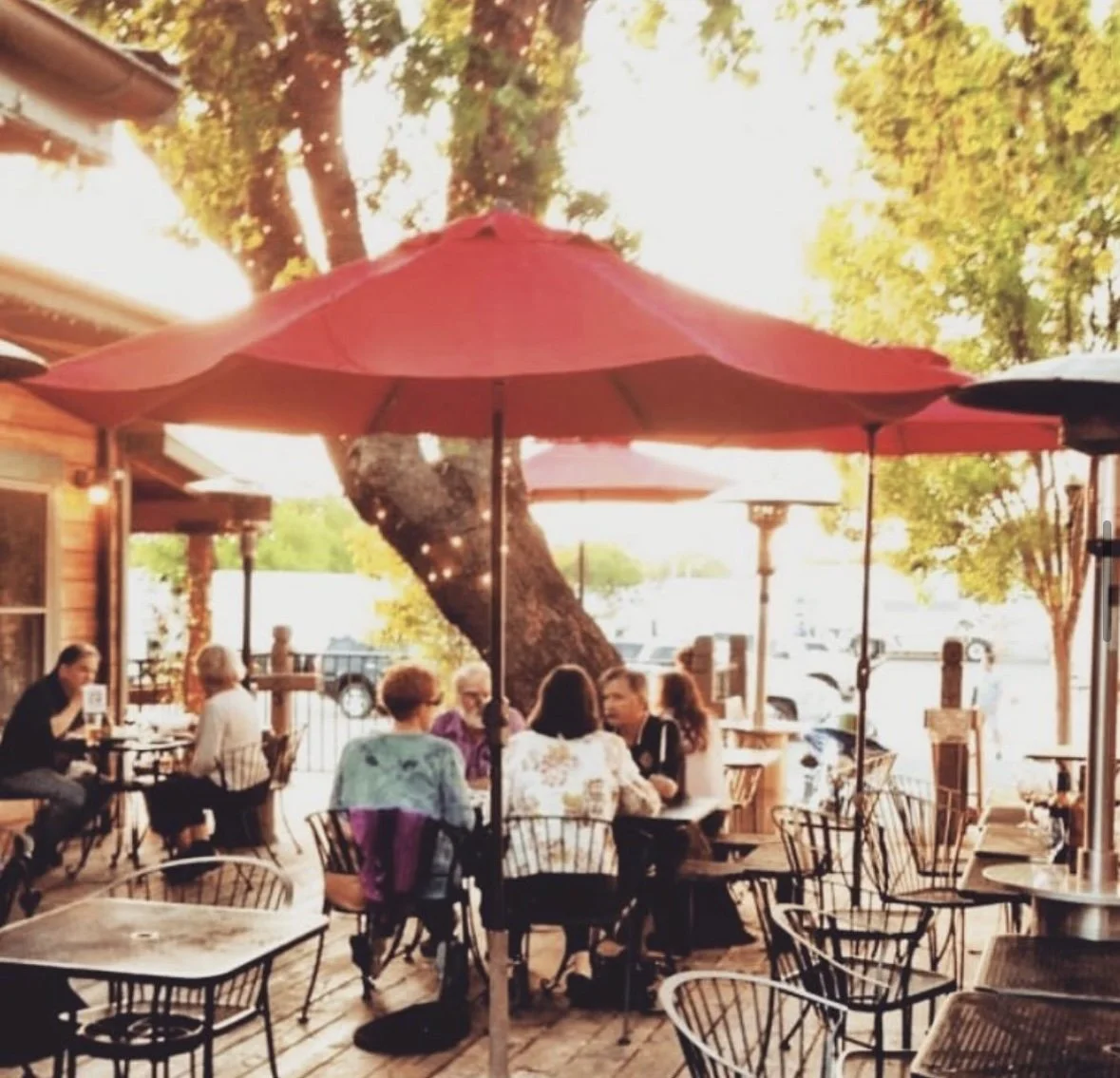 People sitting at outdoor tables under umbrellas at a restaurant or cafe, with trees and parked cars in the background during daylight.