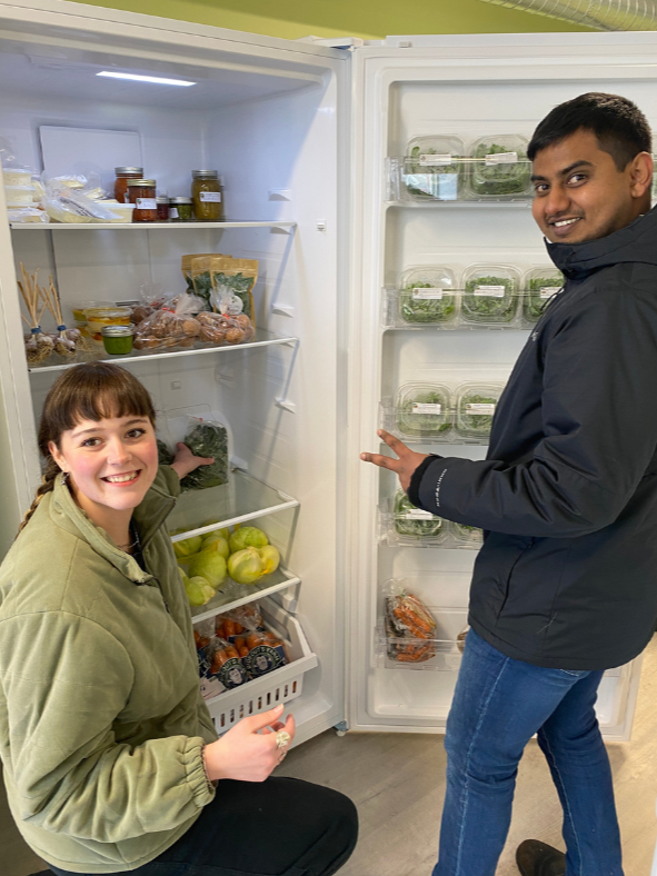 Two people standing in front of an open refrigerator stocked with vegetables and preserves