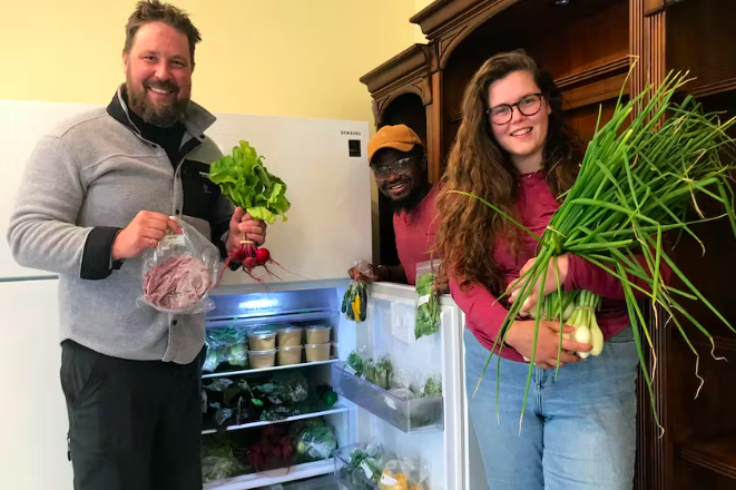 Three people holding up vegetables, meat and herbs and standing around a refrigerator