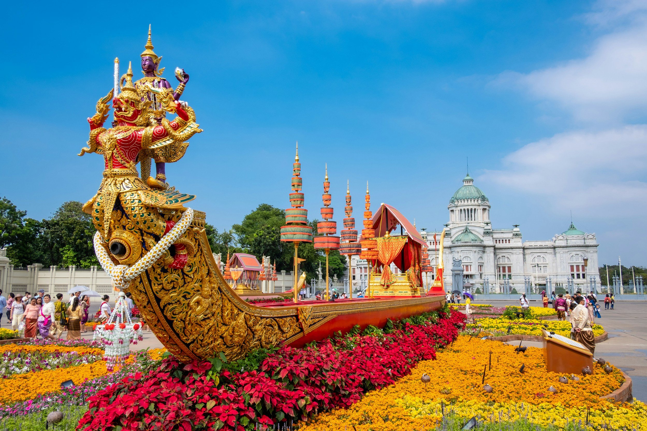 Elaborate Thai royal barge sculpture surrounded by colorful flowers, with a historical European-style building in the background and people walking nearby.