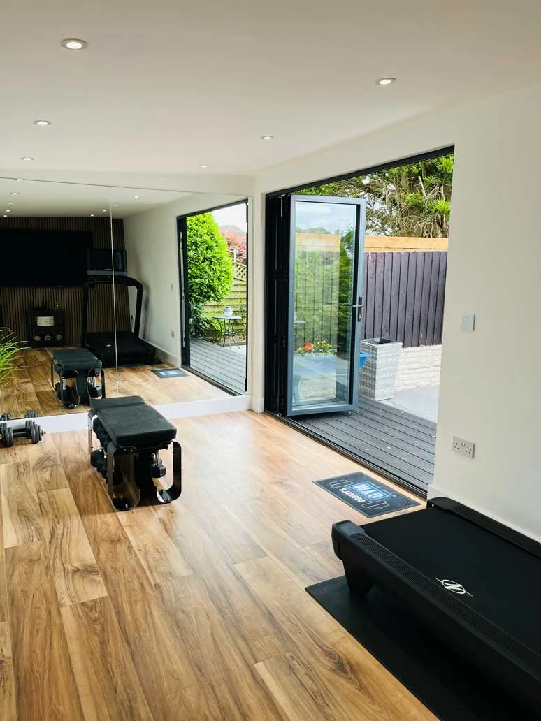 A home gym interior inside a garden building featuring wood-look flooring, large full-length mirrors along one wall, a weight bench with dumbbells, a treadmill near bi-fold glass doors, and recessed ceiling spotlighting.