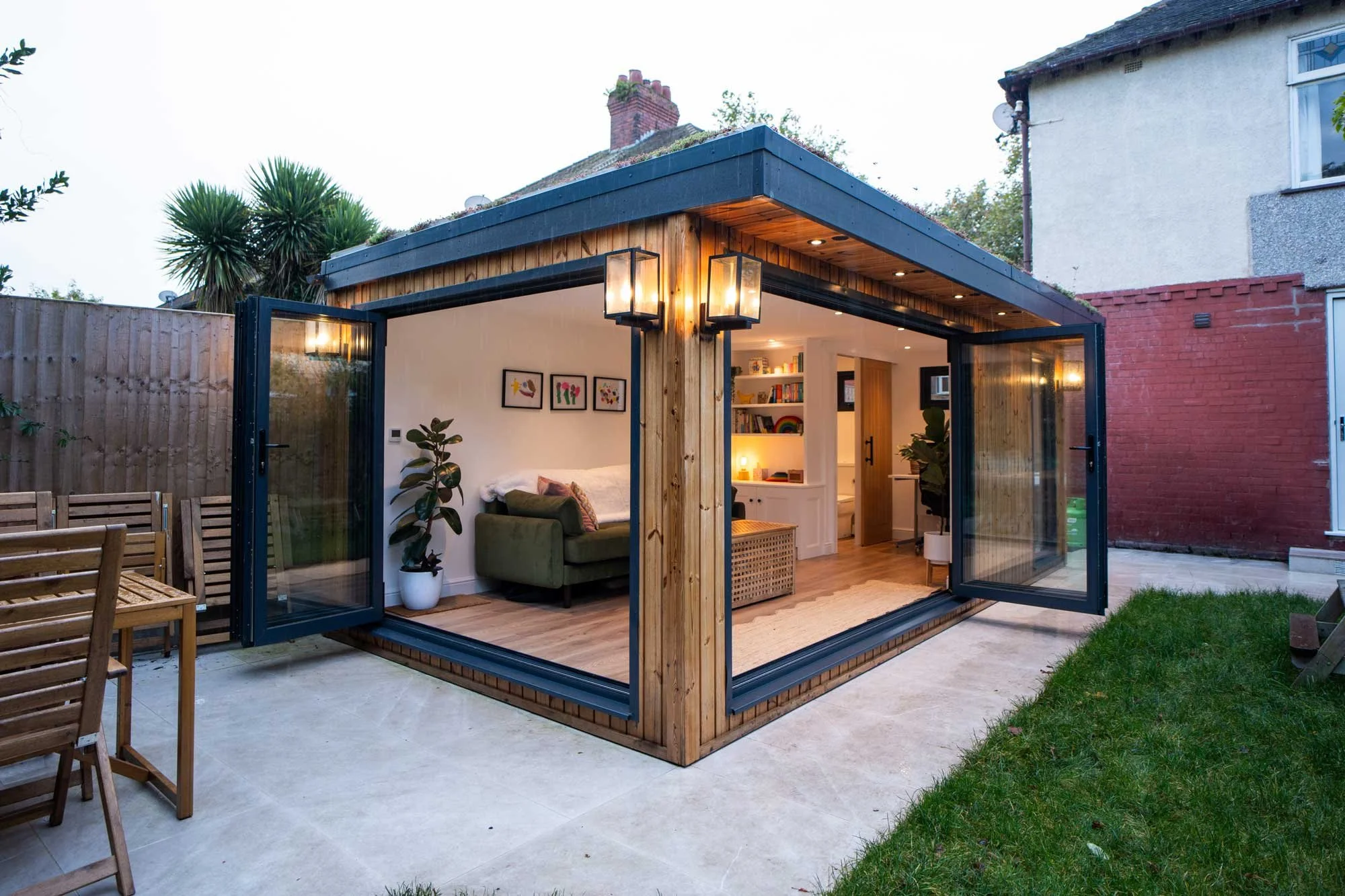 Exterior view of a bespoke wooden garden studio with open black-framed bi-fold doors, a living sedum roof, exterior wall lights, and a cozy interior furnished with an olive green sofa and built-in shelving.