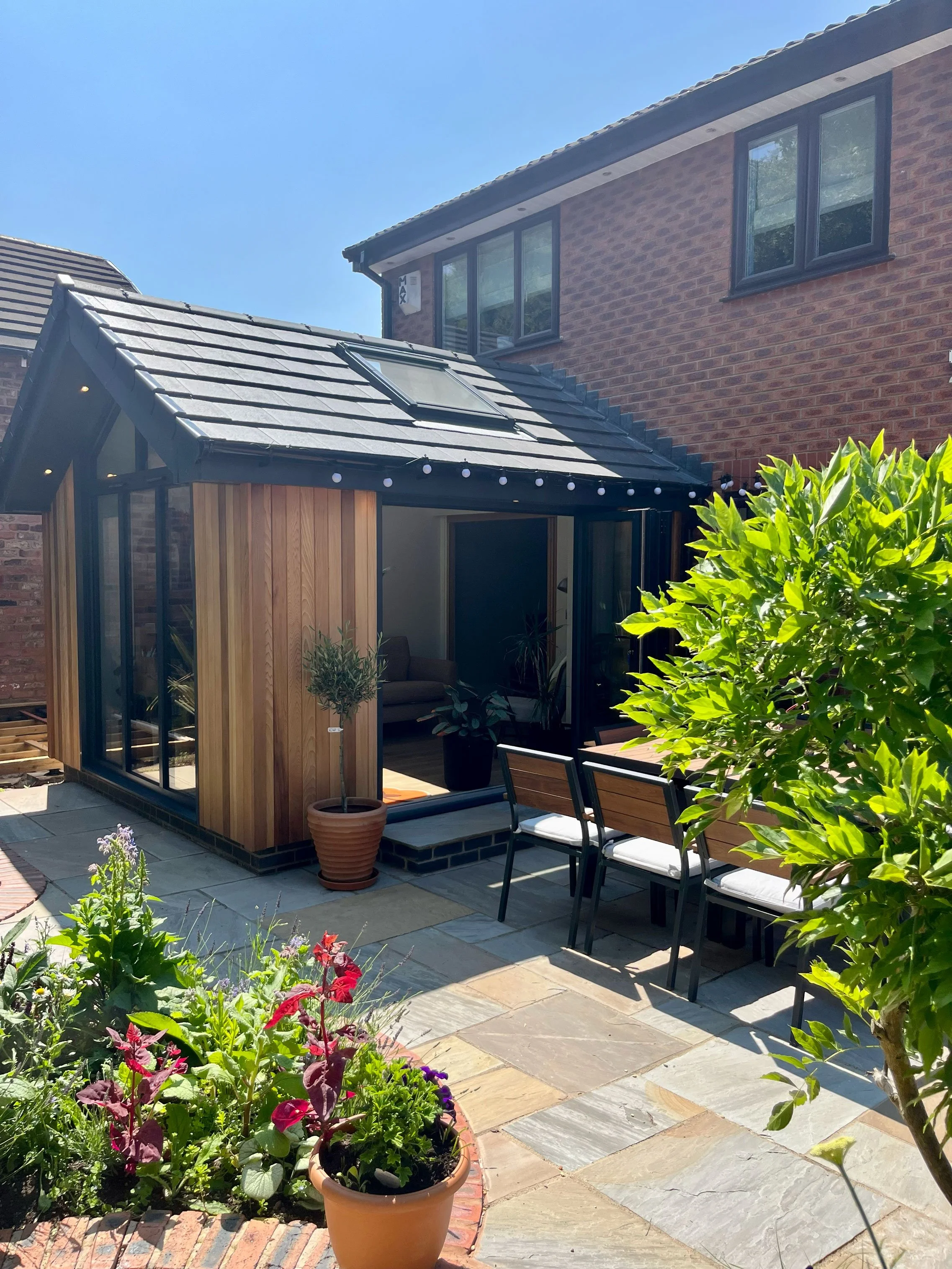 Modern pitched-roof home extension featuring vertical timber cladding, dark slate roof tiles, a skylight, and black-framed glass doors, adjoining a flagstone patio with outdoor dining furniture and garden greenery.