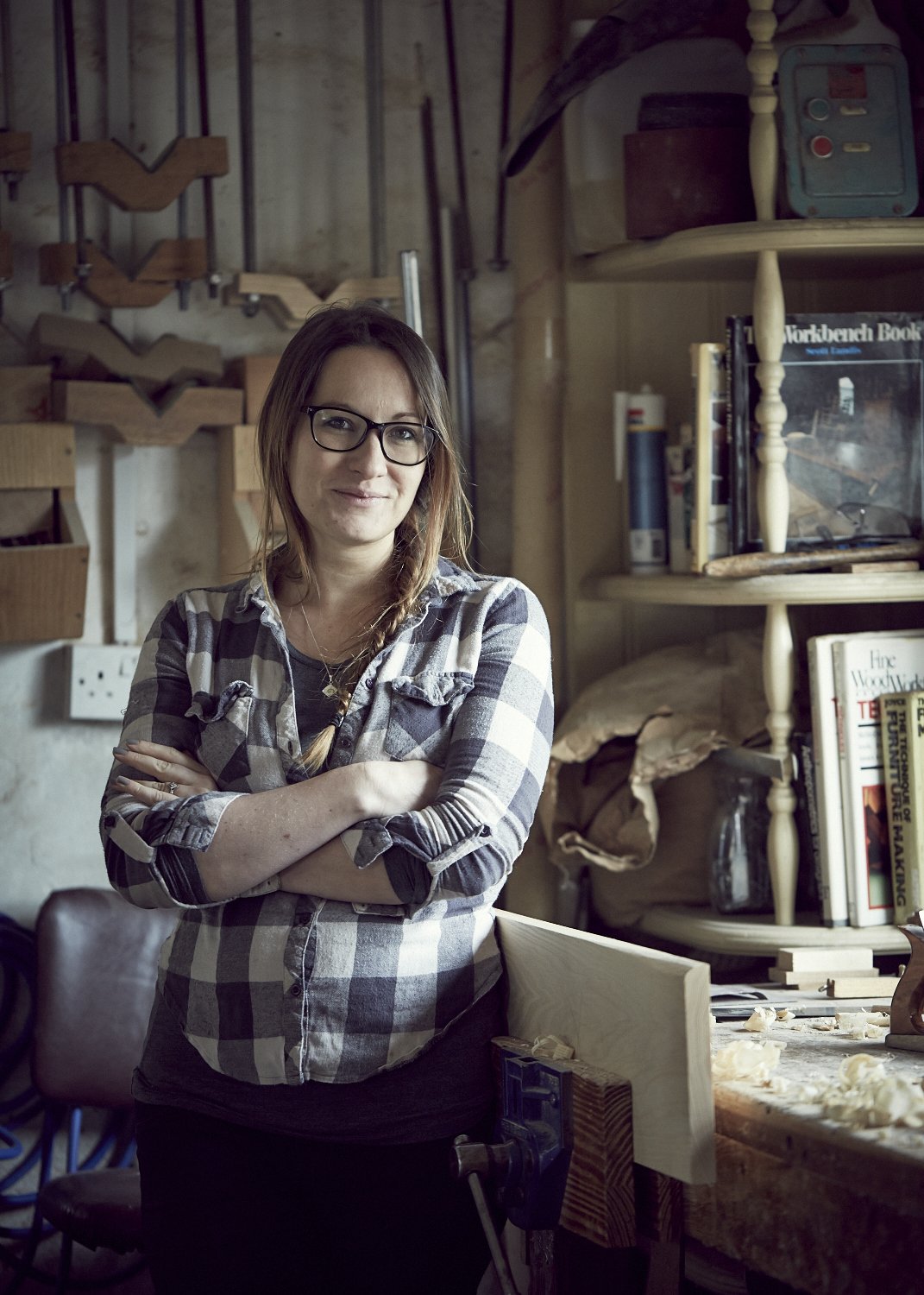 A woman wearing glasses and a black-and-white plaid shirt standing with crossed arms in a woodworking workshop surrounded by tools, wood, and books.
