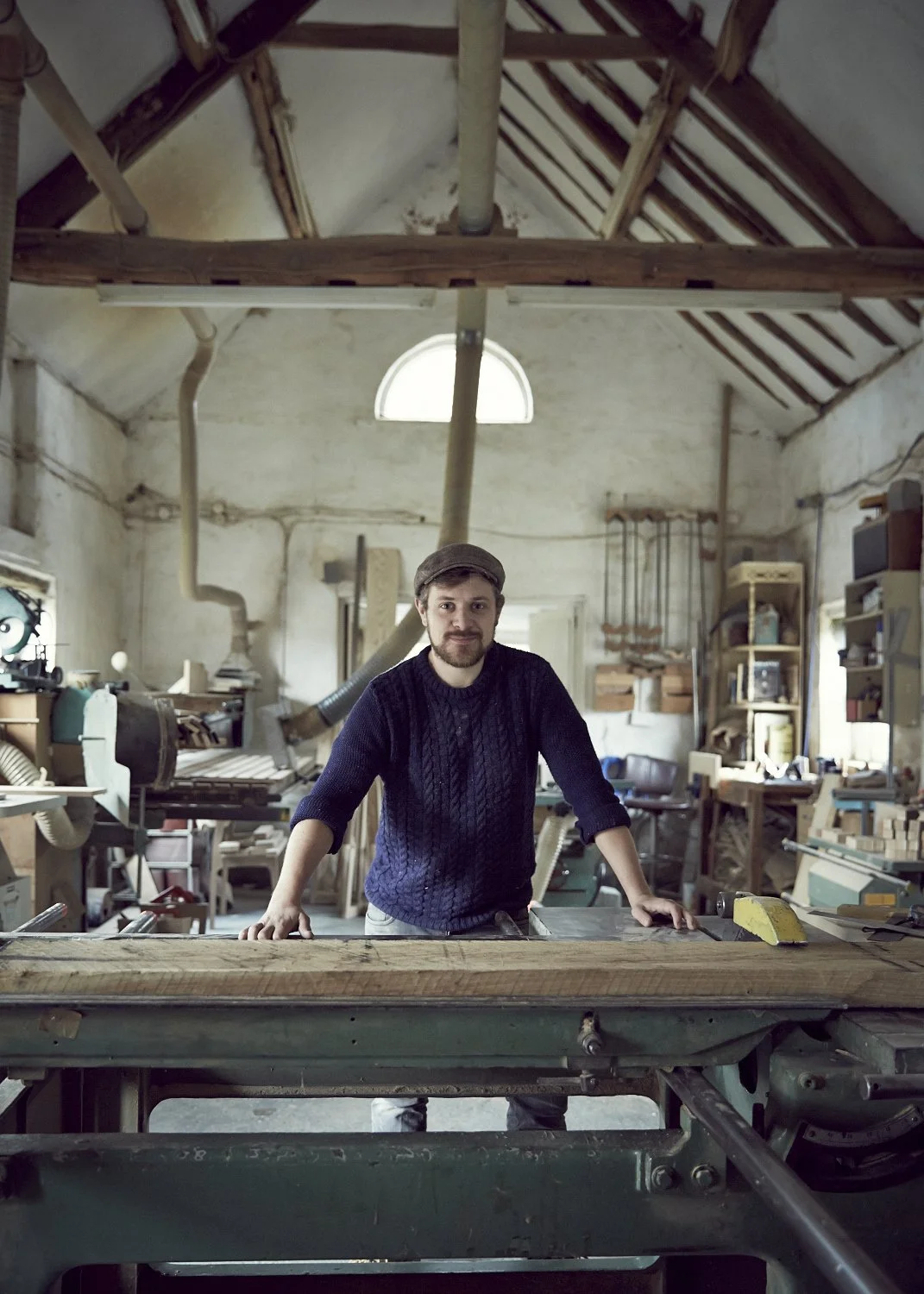 A man in a woodworking shop standing behind a large table saw, surrounded by woodworking tools and shelves filled with pieces of wood.