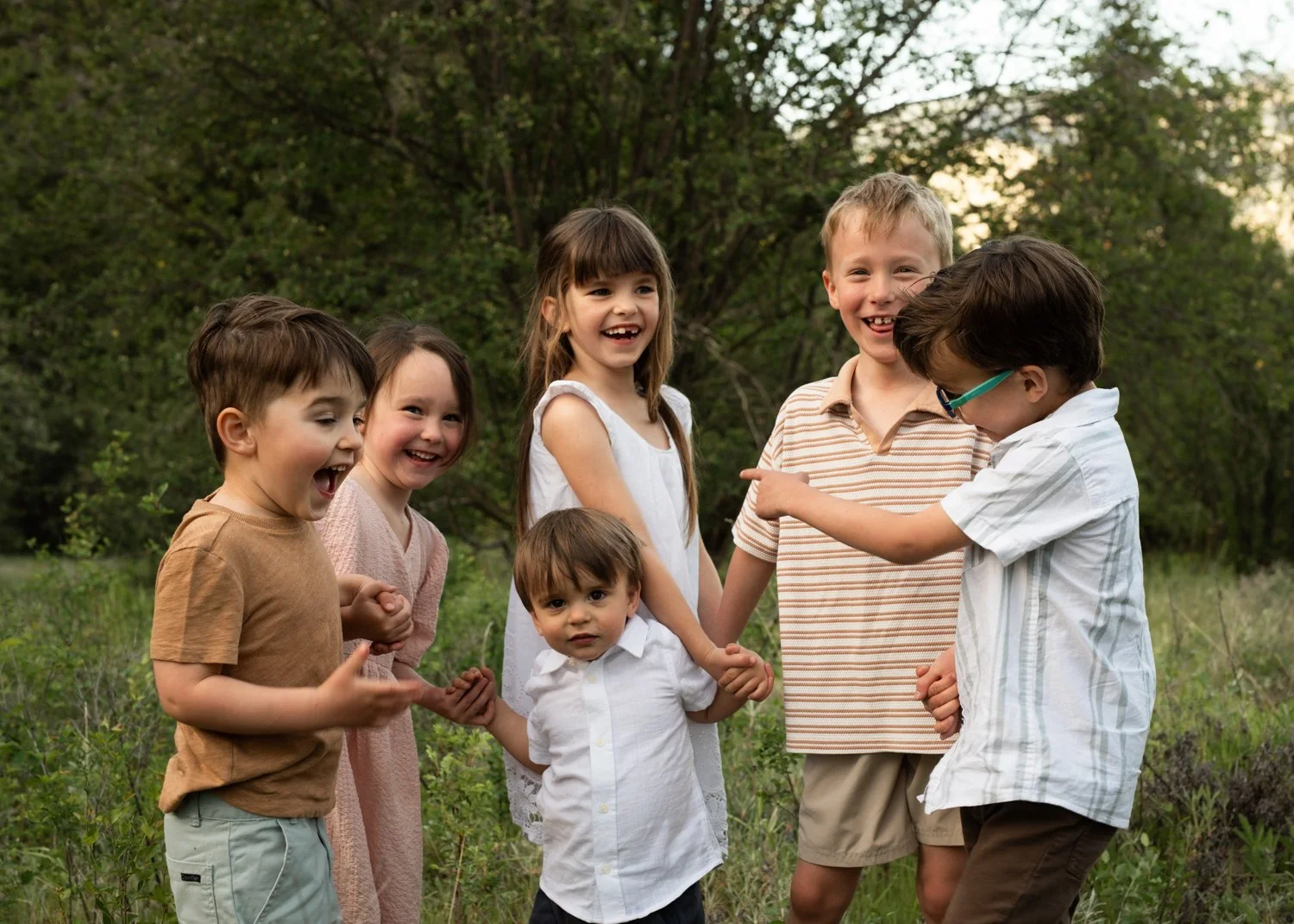 Group of six children playing outdoors together in a grassy area with trees in the background, laughing and smiling.