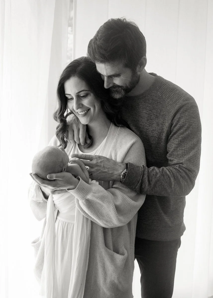 Black-and-white photo of parents snuggled together standing and holding newborn baby beside window with natural lighting