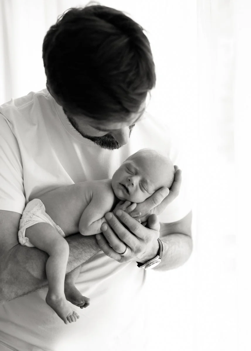 Dad holding newborn on arm with natural light pouring in from window for soft and glowing light