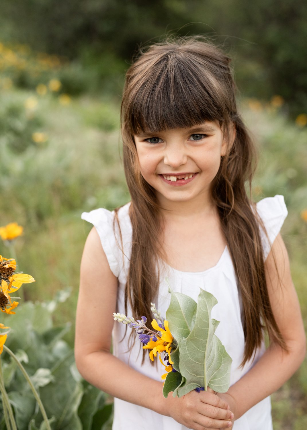 A young girl with long brown hair and a missing front tooth, wearing a white dress, smiling and holding a bouquet of flowers outdoors in a field with yellow flowers.