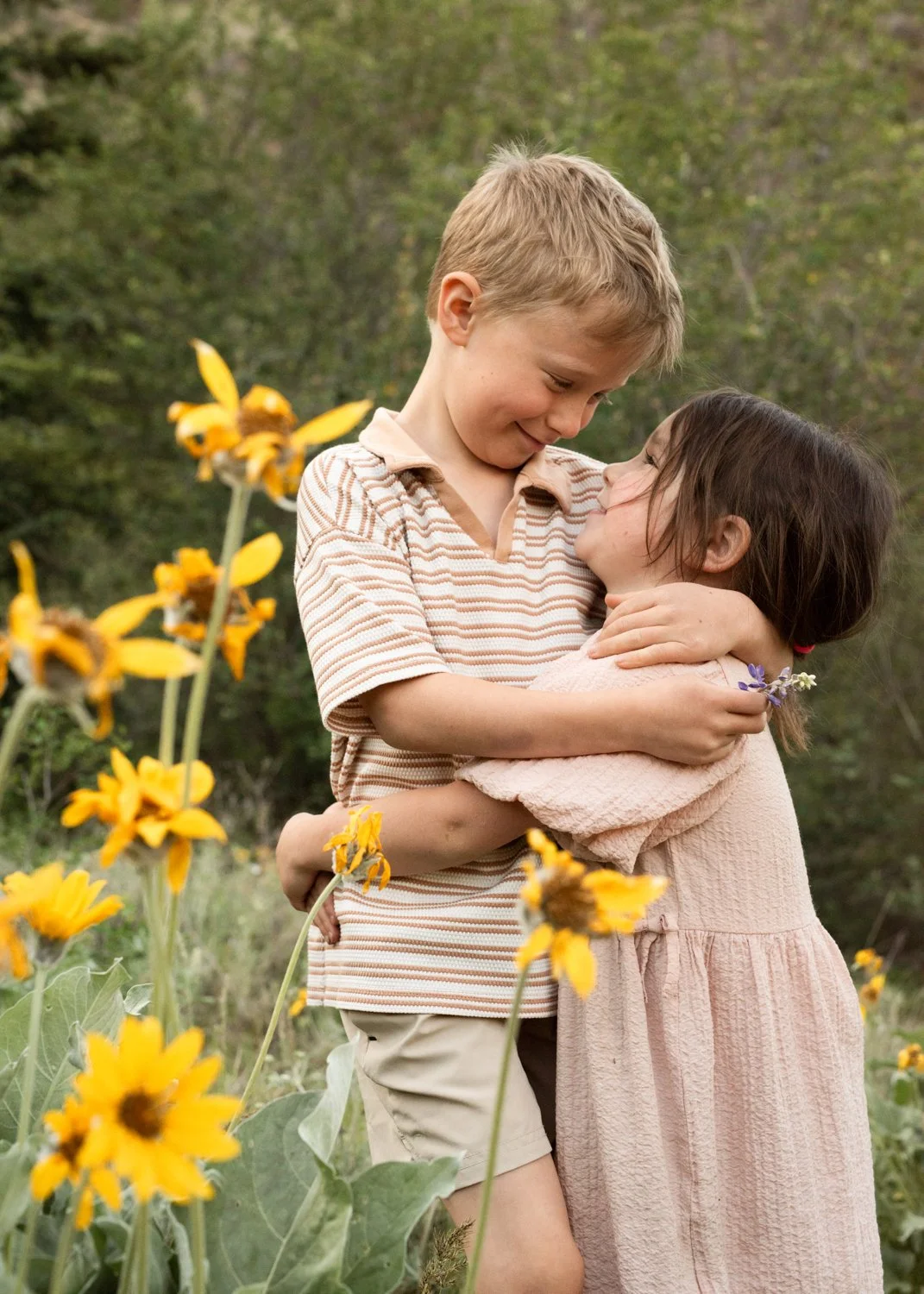 A young boy and girl hugging in a field of yellow flowers with trees in the background, smiling at each other.