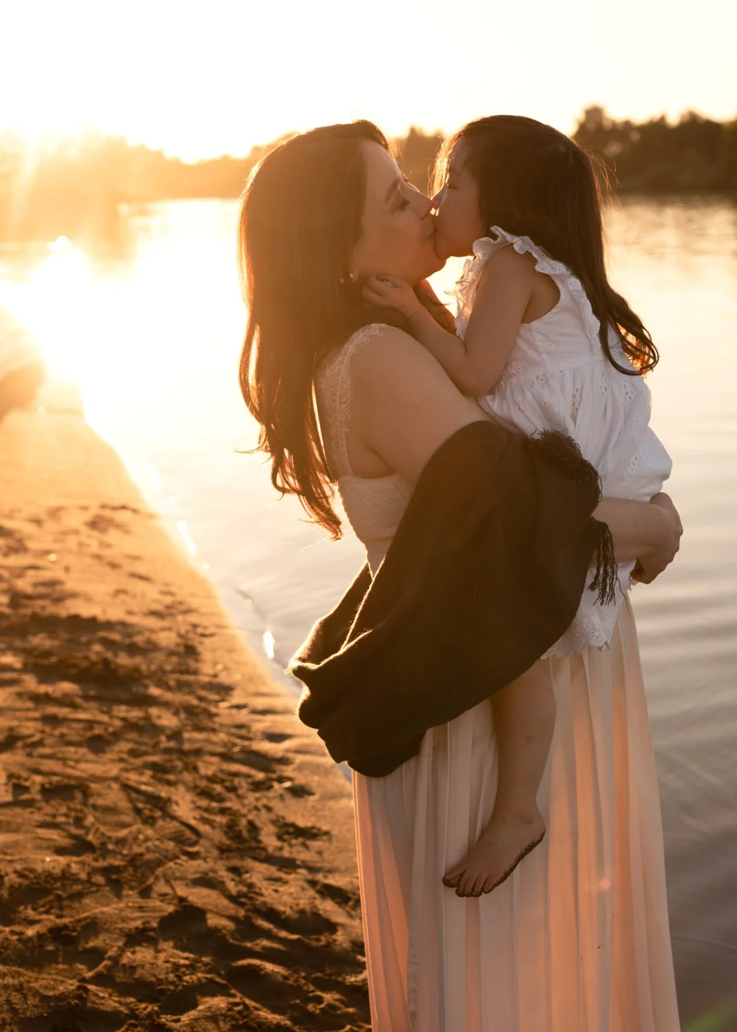 A woman is holding a young girl, and they are kissing by a river at sunset, with the woman's arms around the girl's waist and the girl's feet dangling.