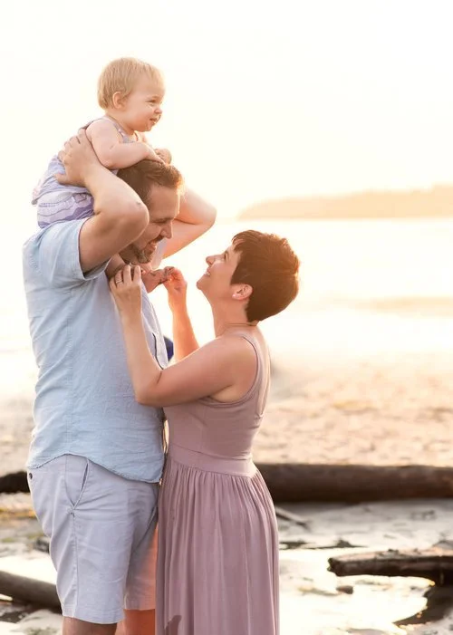 Family of 3 at White Rock beach with Dad holding little girl on his shoulders while mom holds baby's feet and looks up