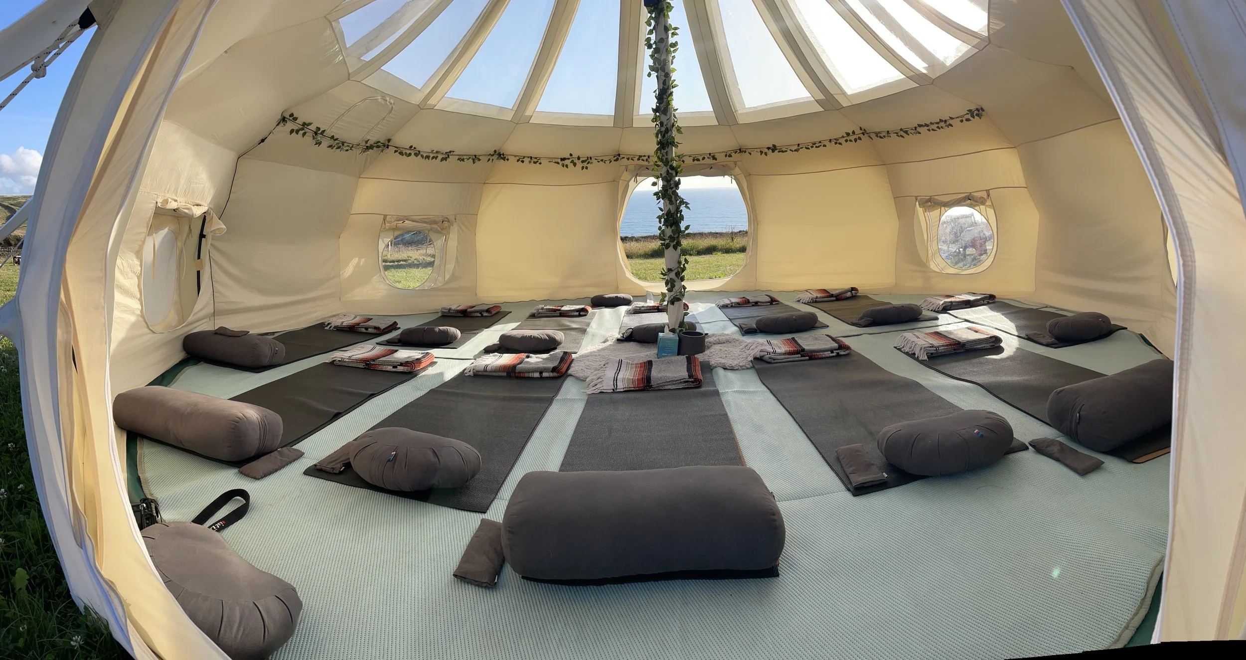 Interior of a large, cream-colored canvas tent with a view of the ocean through multiple windows, featuring black mats and round cushions arranged on the floor, with decorative greenery hanging from the ceiling.