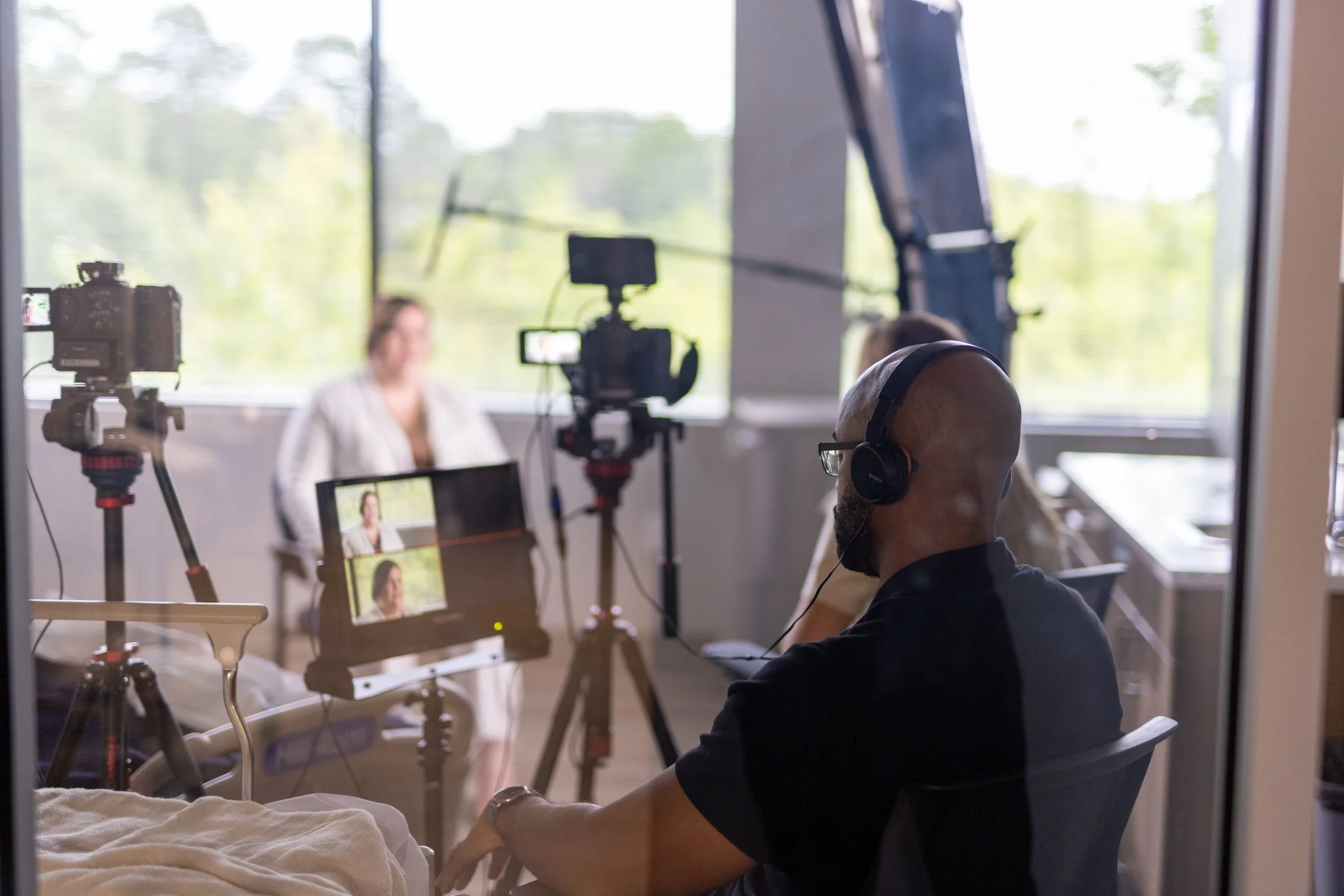A person in a dark shirt and headphones sits at a table in a room with large windows, filming a woman who is seated and speaking in front of a camera. Multiple cameras and equipment are set up facing the woman. Outside the window, greenery is visible.