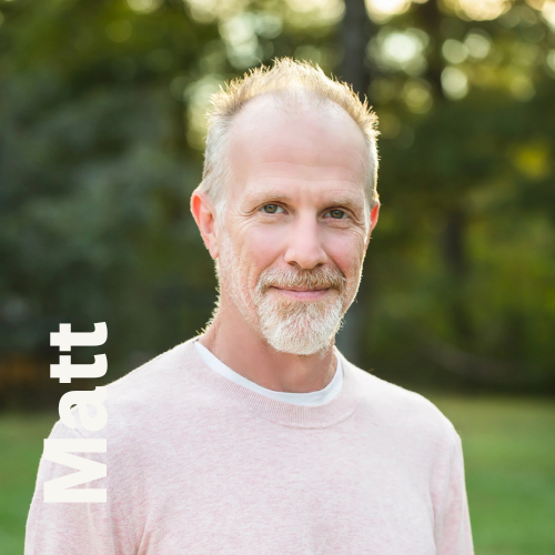 A middle-aged man with a beard and light hair standing outdoors with trees in the background, wearing a light-colored sweater.