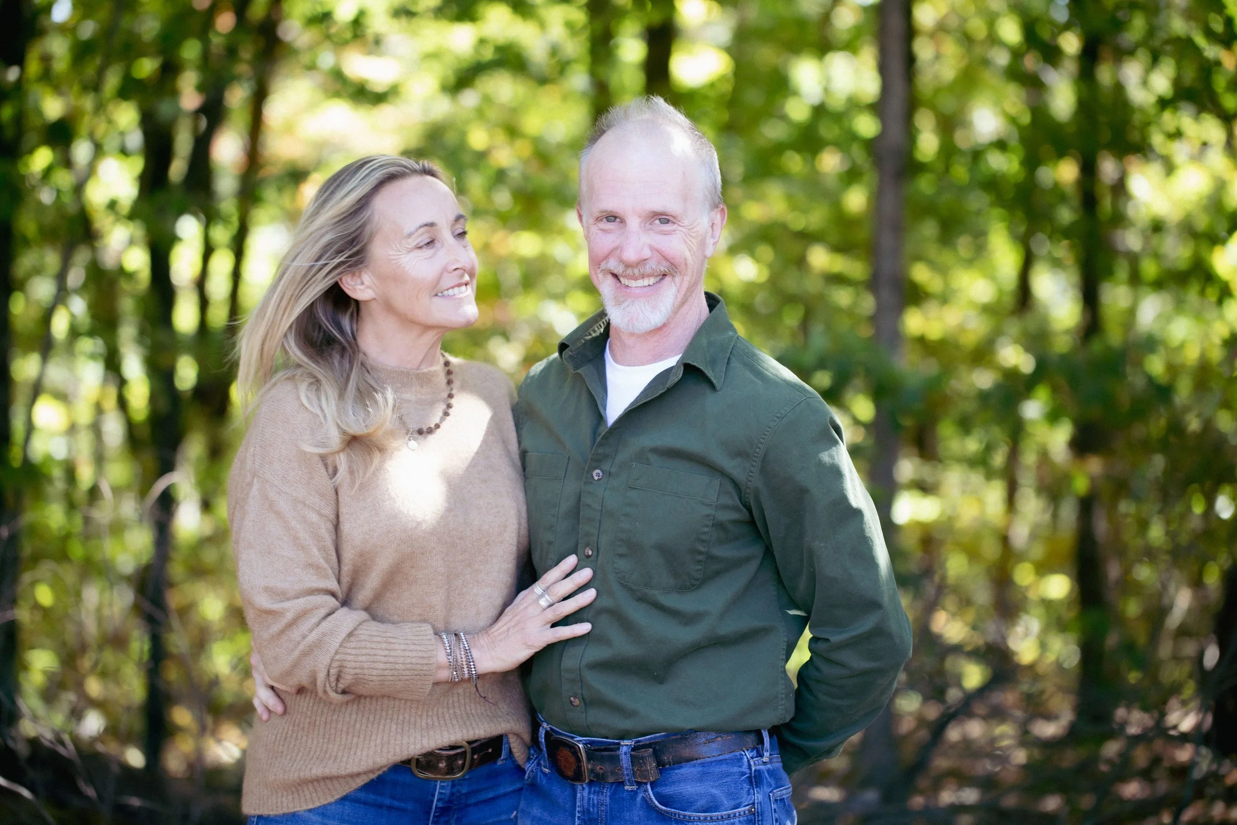 A smiling couple standing outdoors in a wooded area, with the woman touching the man's chest.