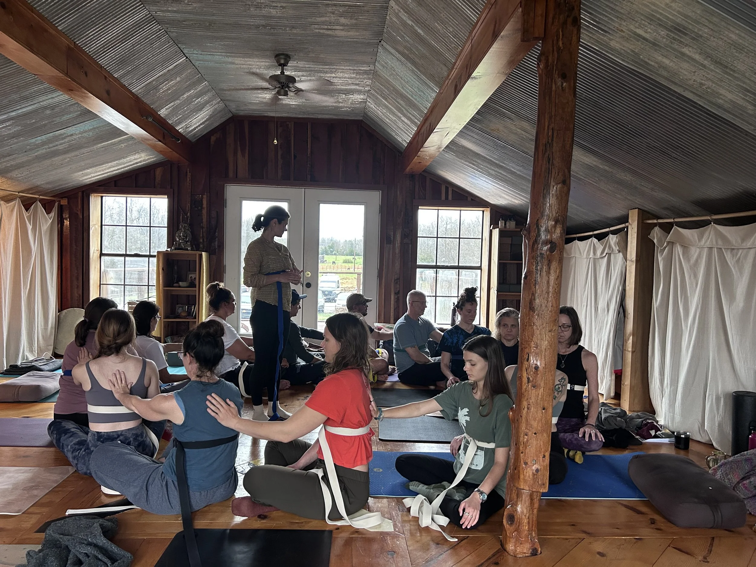 A group of people participating in a yoga or meditation class inside a wooden cabin with large windows, wooden beams, and curtains.