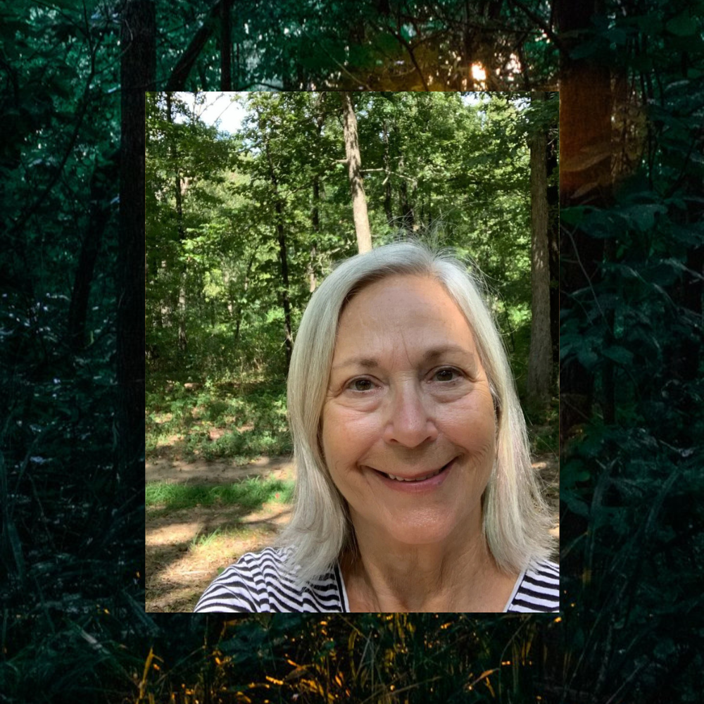 A woman with shoulder-length gray hair smiling in a forested area.