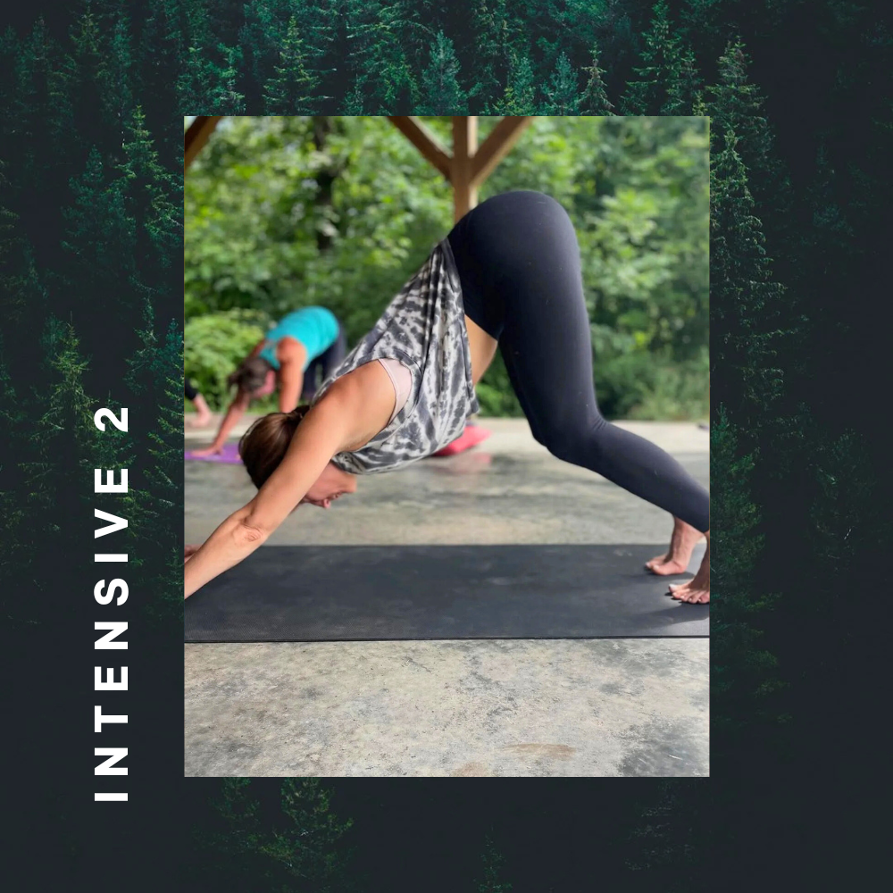 A woman practicing yoga outdoors in a downward dog pose, with her hands and feet on the ground and hips raised, on a black yoga mat surrounded by greenery.