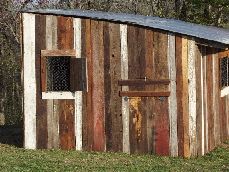 A rustic wooden chicken coop with small windows covered with wire mesh, located outdoors on grassy ground with trees in the background.