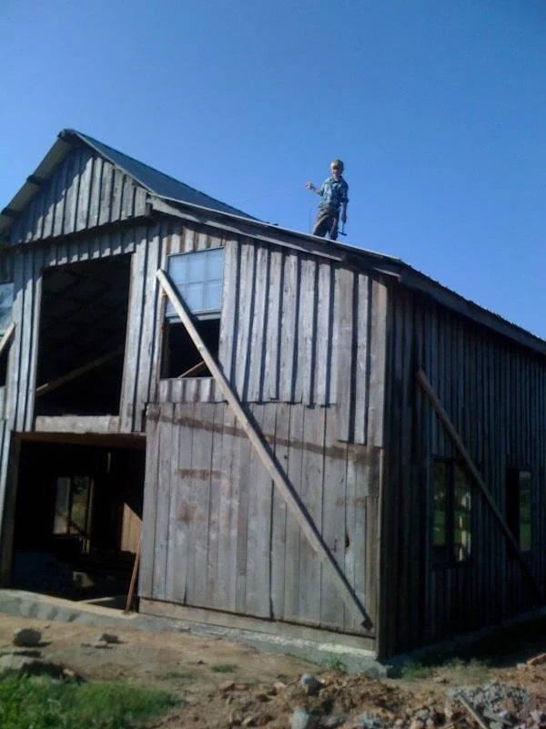 Person standing on roof of a barn under construction, with wooden panels and open gaps visible.